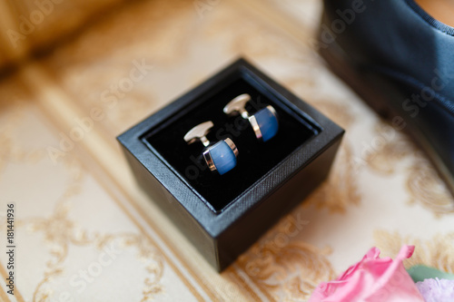 Cufflinks for the groom in the box on the background of the boutonniere with fresh flowers and classical leather shoes , men's accessories of a businessman on a sofa.