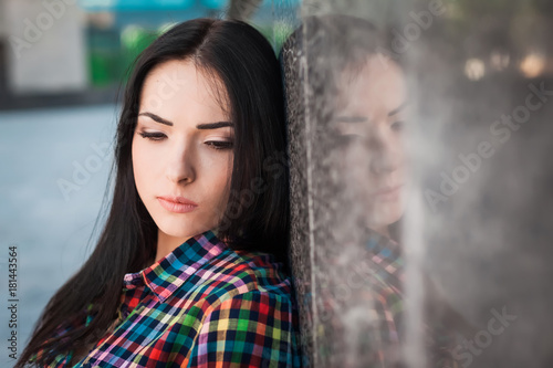 Portrait of young hipster girl with long hair leaning against the wall in the street, wearing colorful shirt in cage on the urban background