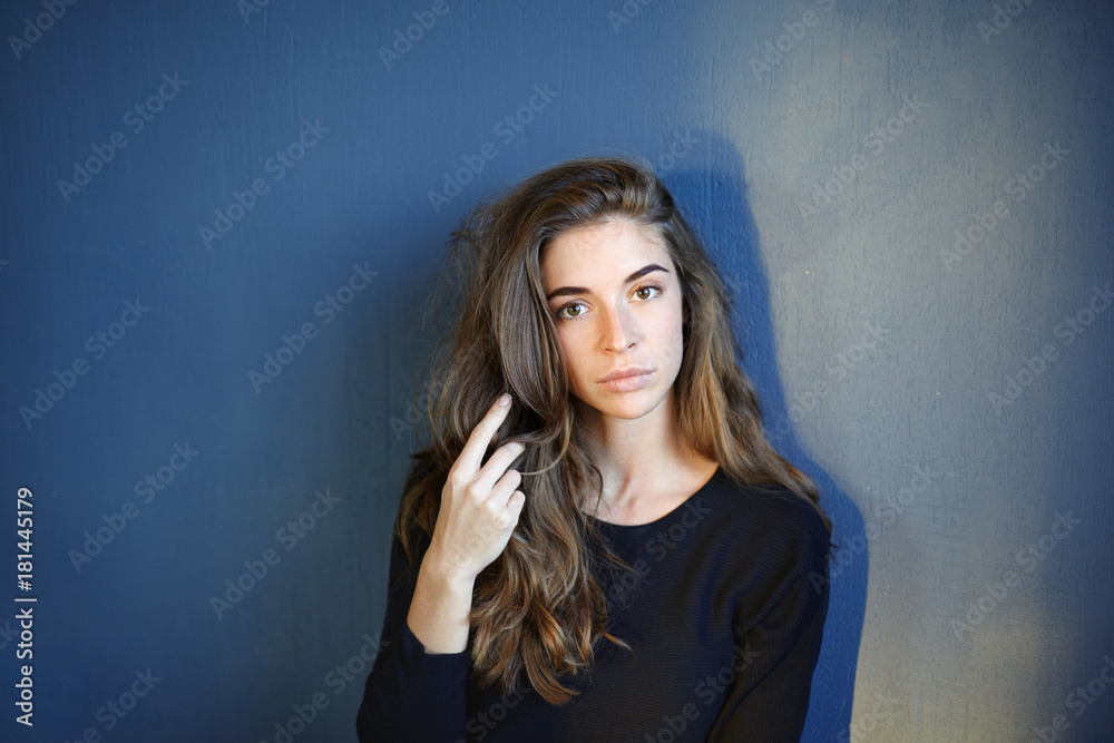 Isolated shot of tender young female ballet dancer, actress or model with voluminous hair having serious look, standing at blank studio wall with copy space for your text or advertising content
