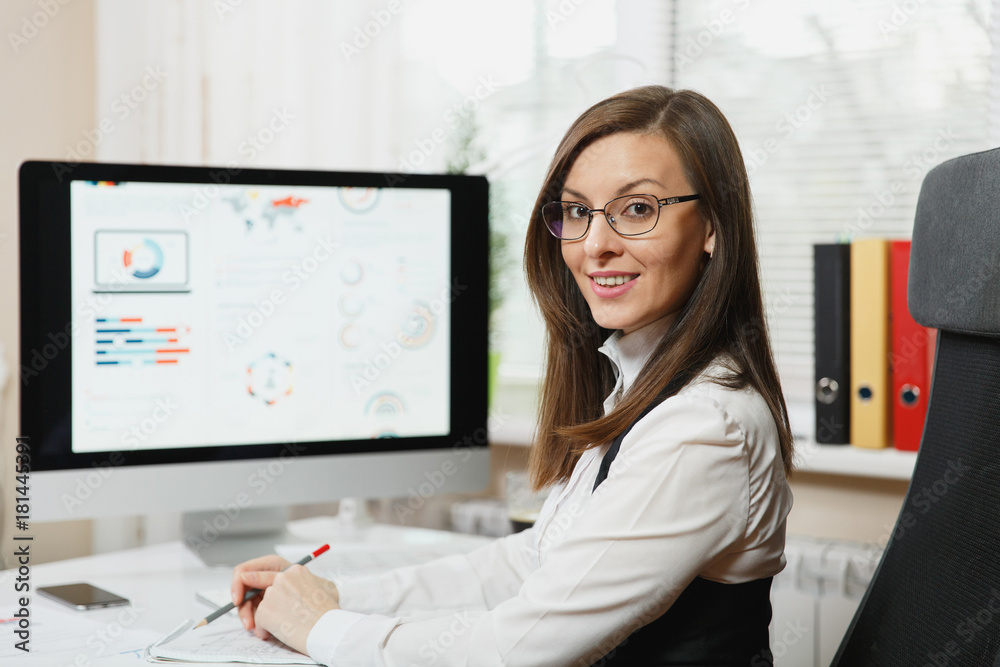 Beautiful smiling brown-hair business woman in suit and glasses working at computer with documents in light office, looking at the camera, against the background of the monitor, close up. Beautiful smiling brown-hair business woman in suit and glasses working at computer with documents in light office, looking at the camera, against the background of the monitor, close up.