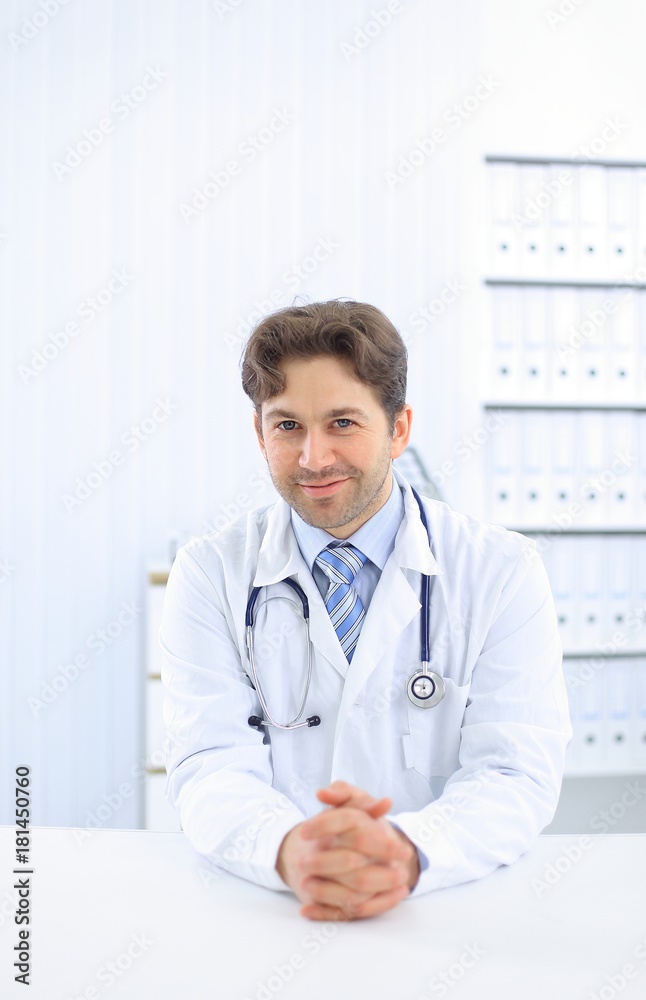 Handsome young doctor in white coat is looking at camera and smiling while standing in office