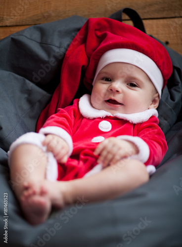 Baby girl in Santa Claus costume sits among pillow and smiles.