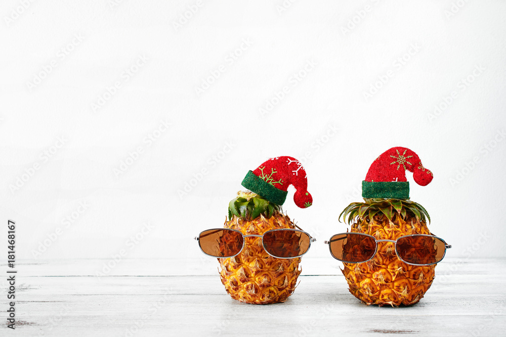 Closeup of two pineapples in sunglasses and christmas hat over white background with copy space. Christmas in exotic country concept. Closeup of two pineapples in sunglasses and christmas hat over white background with copy space. Christmas in exotic country concept.