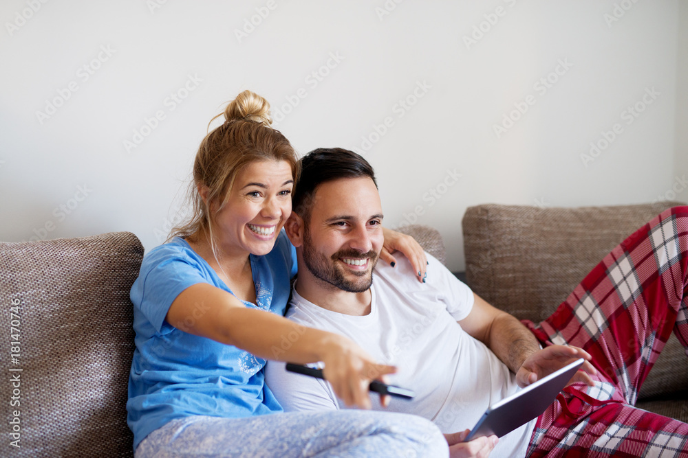 Smiling happy beautiful girl showing with a finger on tv to her handsome boyfriend while lying with a tablet next to her. Smiling happy beautiful girl showing with a finger on tv to her handsome boyfriend while lying with a tablet next to her.