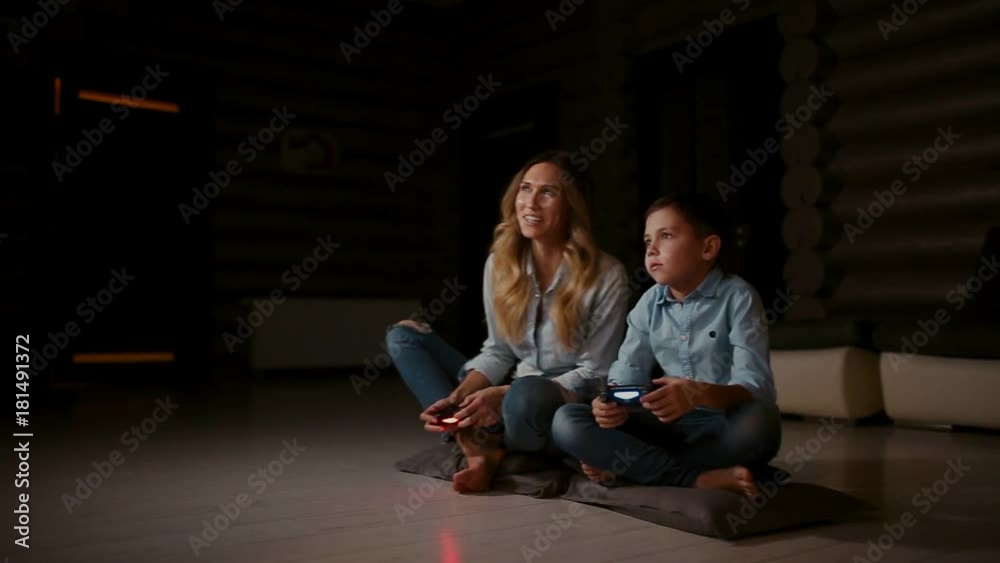 Beautiful mother with her son playing video games on gaming console in the living room of his house. Happy family in a country house. Beautiful mother with her son playing video games on gaming console in the living room of his house. Happy family in a country house.