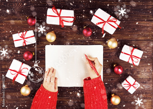 Female hands writing christmas gift list in notebook on wooden background with gifts and red and gold boiis. Toned picture with snow effect. photo