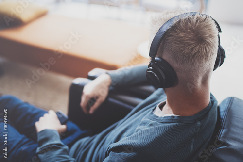 Young man wearing casual clothes,sitting on comfort armchair and listening music in headphones on smartphone at modern home studio.Horizontal.Blurred background.