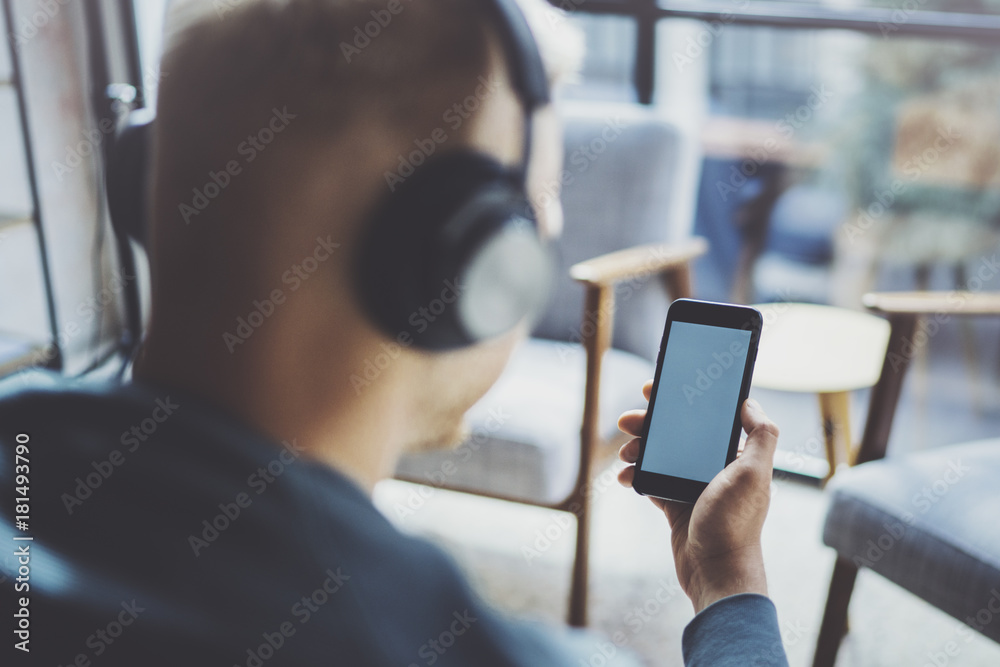 Young man wearing eye glasses and casual clothes listening music in headphones on smartphone at modern place.Horizontal mockup.Blurred background.