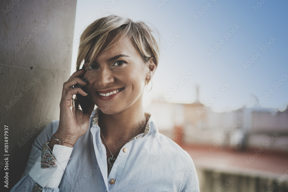 Portrait of smiling businesswoman talking with partner on smartphone device while working remotely in business travel.Attractive woman owner communicating on cellular while looking at camera.Blurred. Portrait of smiling businesswoman talking with partner on smartphone device while working remotely in business travel.Attractive woman owner communicating on cellular while looking at camera.Blurred.