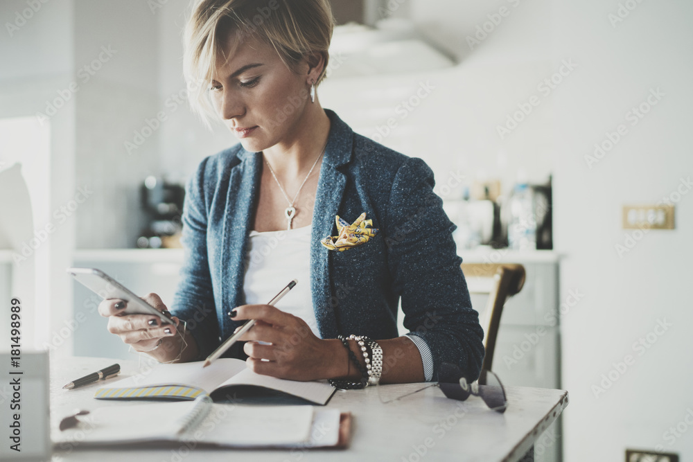Attractive businesswoman texting sms with partner via modern telephone while writing some information in note book while sitting at living room in hotel.Blurred background. Attractive businesswoman texting sms with partner via modern telephone while writing some information in note book while sitting at living room in hotel.Blurred background.
