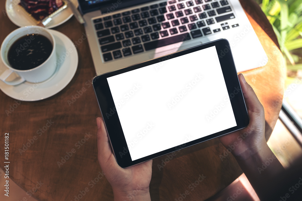 Mockup image of hands holding black tablet pc with blank white screen , laptop , coffee cup and cake on wooden table in cafe Mockup image of hands holding black tablet pc with blank white screen , laptop , coffee cup and cake on wooden table in cafe