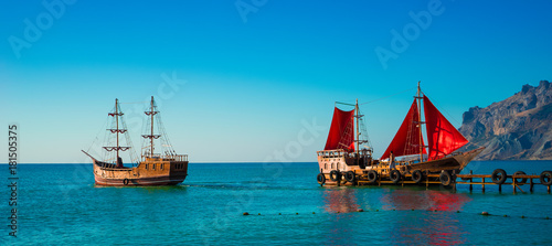 Two old wooden ships. One of them with red sails is on the pier. Scarlet sails, as in the famous story of Alexander Green. The second sails from the pier without raised sails. Koktebel, Crimea.