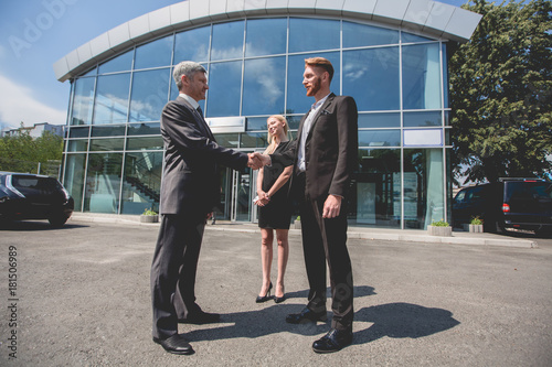 Young man and woman buying electro car. Family couple and cae dealer in front of car showroom  men shaking hands.