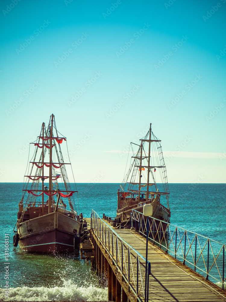 Two wooden sailing ships at the wooden pier against the blue sea on a bright sunny afternoon.