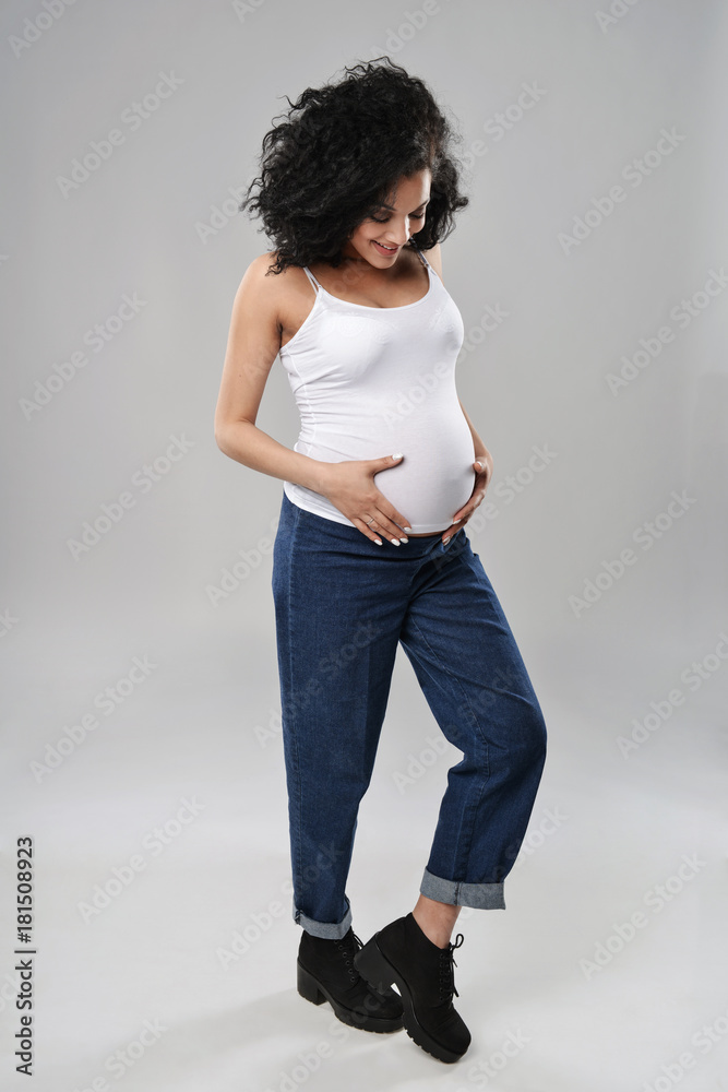 Pregnant woman standing in full length in jeans and white top, smiling looking down at her belly, over grey background Pregnant woman standing in full length in jeans and white top, smiling looking down at her belly, over grey background