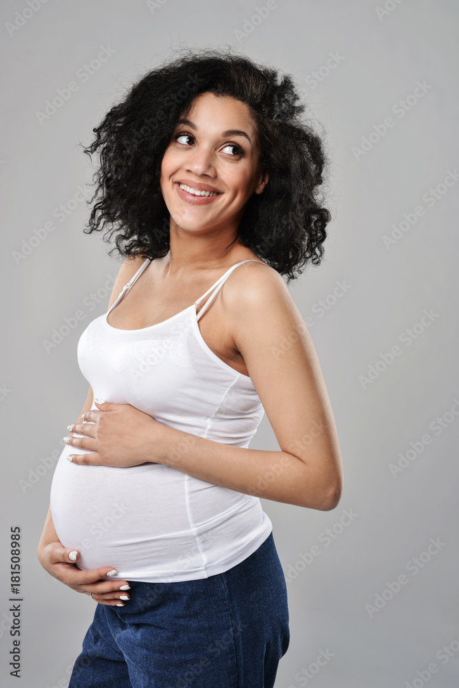 Pregnant woman smiling looking away out of frame with hands on tummy, over grey background Pregnant woman smiling looking away out of frame with hands on tummy, over grey background
