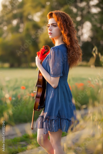 Beautiful romantic girl with red hair and blue dress holding violin on nature field of poppy flowers. Photo of sensual woman