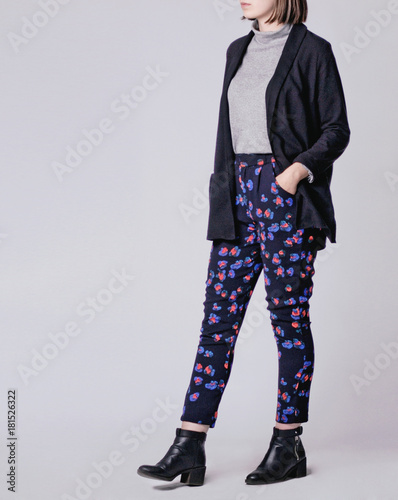 Woman wearing stylish smart outfit with black patterned floral cigarette trousers, gray turtleneck, black oversized blazer and black ankle boots isolated on gray background. Copy space