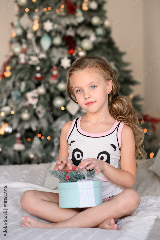 Christmas celebration. Beautiful little girl sitting on a bed  near the Christmas tree. Happy little smiling girl with christmas gift box. Christmas miracles. Happy New Year