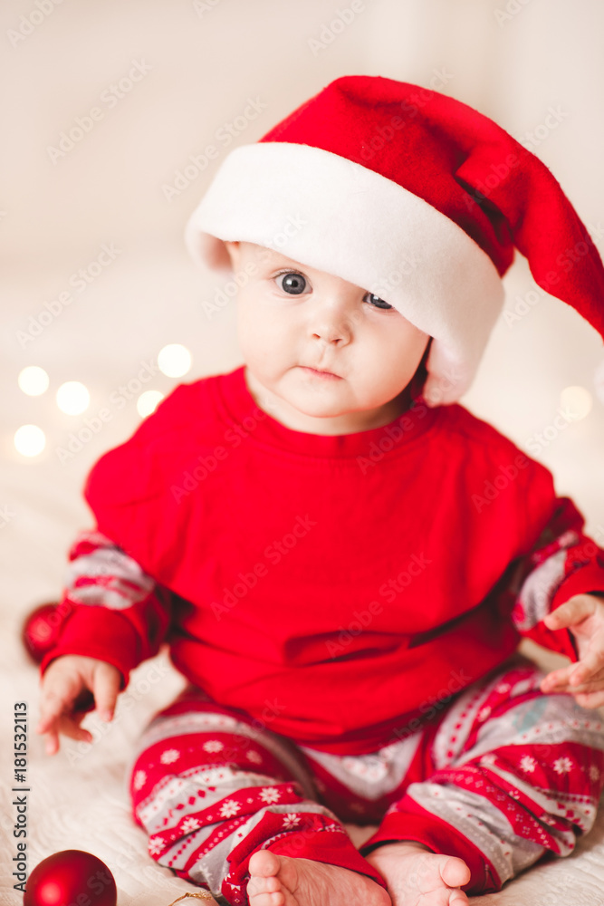 Cute baby sitting in bed wearing santa claus hat and suit. Looking at camera. Holiday season. Childhood. Cute baby sitting in bed wearing santa claus hat and suit. Looking at camera. Holiday season. Childhood.