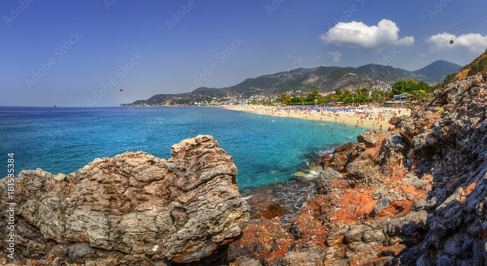 Seascape with rocky mountains and blue sea on sunny summer day. Panoramic landscape of Alanya beach, Turkey. Summer vacation on tropical resort. Paradise lagoon bay with tourists on beach.