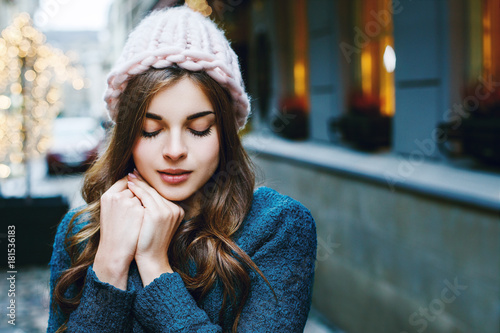 Outdoor close up portrait of young beautiful girl with long hair wearing pink big loop hat, blue sweater posing in street of european city. Christmas, winter holidays concept. Copy, empty space 