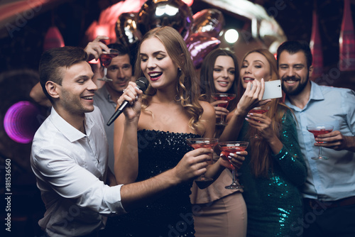 A woman in a black dress is singing songs with her friends at a karaoke club. Her friends do selfie.