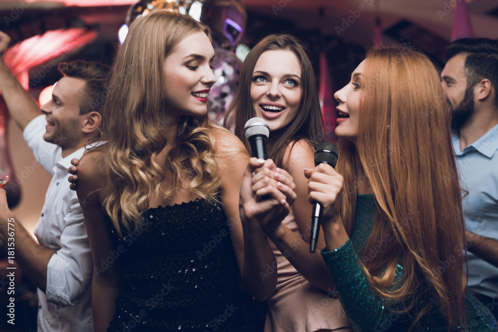 Three beautiful girls sing in a karaoke club. Behind them are men waiting for their turn.