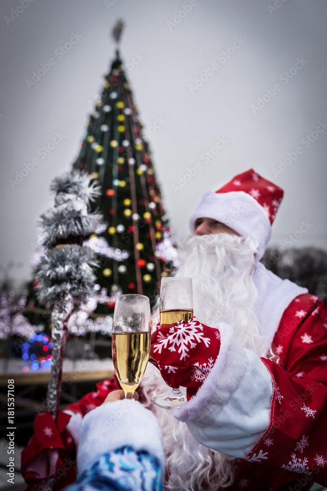 Obraz premium Celebration and New Year concept. Man with beard holds champagne glass. Guy near Christmas tree in the park. Santa Claus in red hat with cheerful face. Happy new Year! Cheers!