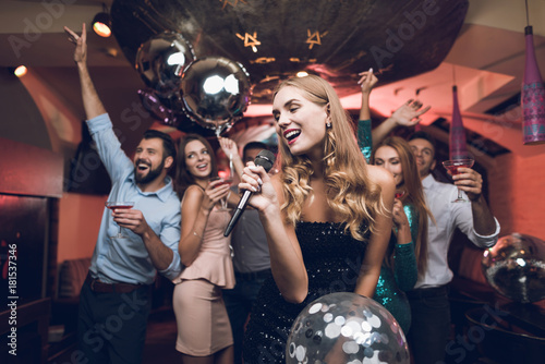 Young people have fun in a nightclub and sing in karaoke. In the foreground there is a woman in a black dress.