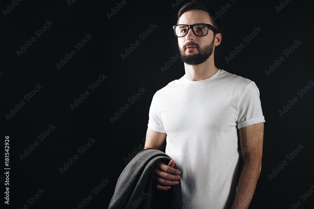 Handsome bearded man in glasses posing in a white blank tshirt and holding a coat in his hand background on black wall Handsome bearded man in glasses posing in a white blank tshirt and holding a coat in his hand background on black wall