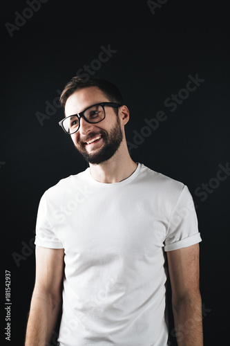 Happy and smiling young bearded man wearing glasses and white blank t-shirt on a dark background. Laughing hipster in an empty tshirt
