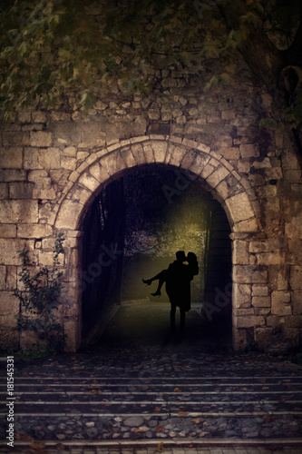 Canvastavla Silhouette of a couple, man holding a woman in his arms, walking through dark arch with yellow backlight