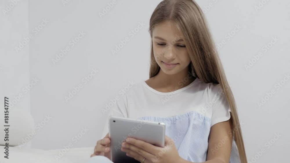 Cute girl sitting on sofa with tablet computer doing homework and playing games