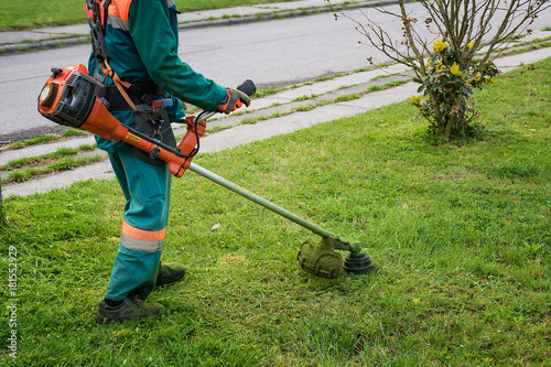 Man in overall and safety helmet trims overgrown grass by grass cutter