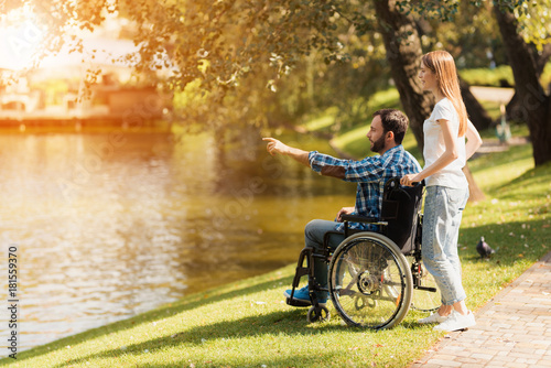 A woman is walking in the park with a man in a wheelchair. They are on the shore of a lake.