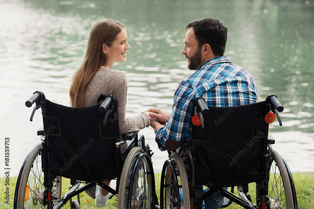 A man and a woman in wheelchairs are sitting on the lake shore. Before them a beautiful lake. They sit hand in hand.