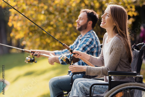 A woman and a man in wheelchairs are sitting on the lake shore. They have spinners in their hands and they are fishing.