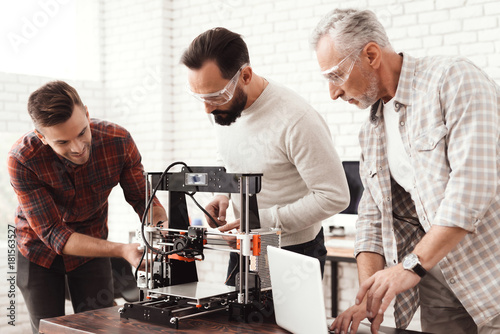 Three men set up a self-made 3d printer to print the workpiece. An elderly man with a laptop is watching his colleagues.