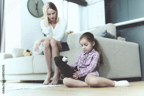The woman is talking to a small offended girl, who sits next to the sofa and holds a toy in her hands.