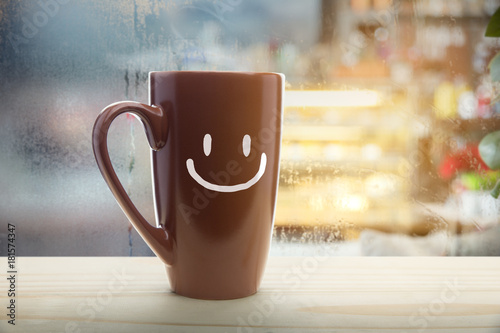 Brown mug of coffee with a happy smile, Steaming red coffee cup on a rainy day window background, Good morning or have a happy day message concept