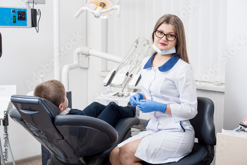 Female dentists examining and working on boy patient. Kid boy in dentist s chair. Doctor looks at camera. Dentist s office