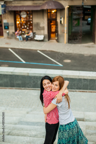 Two young happy sisters hugging in the street. one girl with long brunette hair in red plaid shirt and black trousers, another redhead girl in gray shirt and blue skirt. concept of sincere friendship