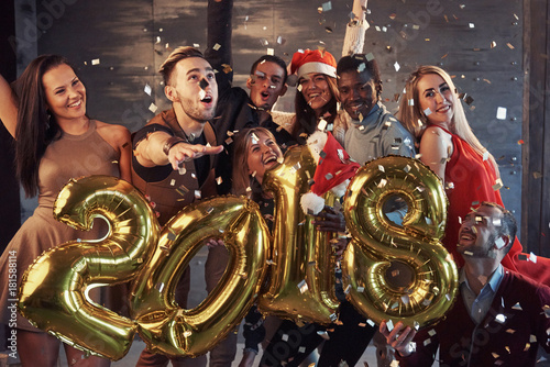 New 2018 Year is coming! Group of cheerful young multiethnic people in Santa hats carrying gold colored numbers and throwing confetti on the party