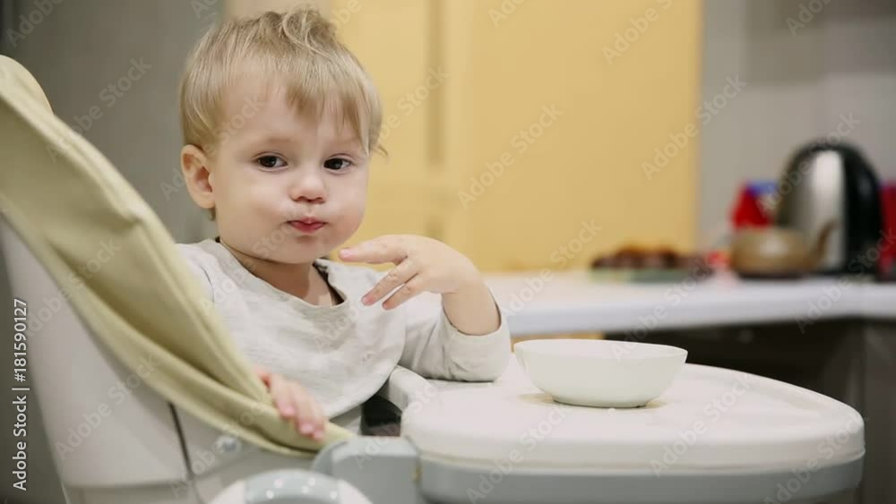 Little boy sitting in the child seat in the kitchen and from the plates is very tasty food. Smiles and plays. Little boy sitting in the child seat in the kitchen and from the plates is very tasty food. Smiles and plays.