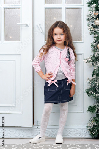 A nice girl of seven with chestnut hair, stands against the backdrop of folding doors decorated in Christmas style, in a blue skirt - a pink jacket. The concept of a holiday