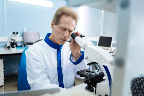 Gene expert. Attractive serious researcher of middle age wearing a uniform and looking into a microscope while making an analysis