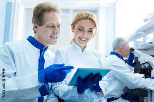 This is wonderful. Beautiful blond young promising researcher holding a tablet and looking at the screen while sitting next to a handsome smiling male scientist and one more biologist working