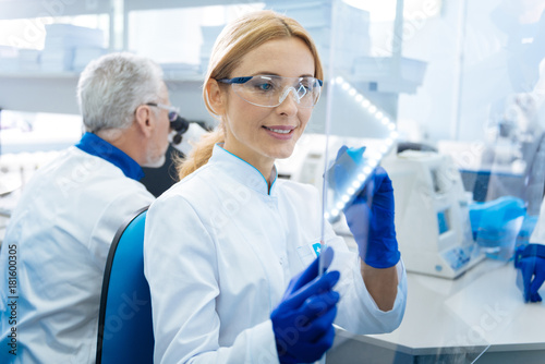 Preparation for examination. Pretty gleeful smiling woman scientist wearing a uniform and glasses and holding a marker and glass while being in the lab