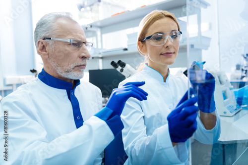 Testing. Attractive gleeful promising scientist wearing a uniform and glasses and holding a marker and glass in the lab and a serious grey-haired researcher sitting next to her